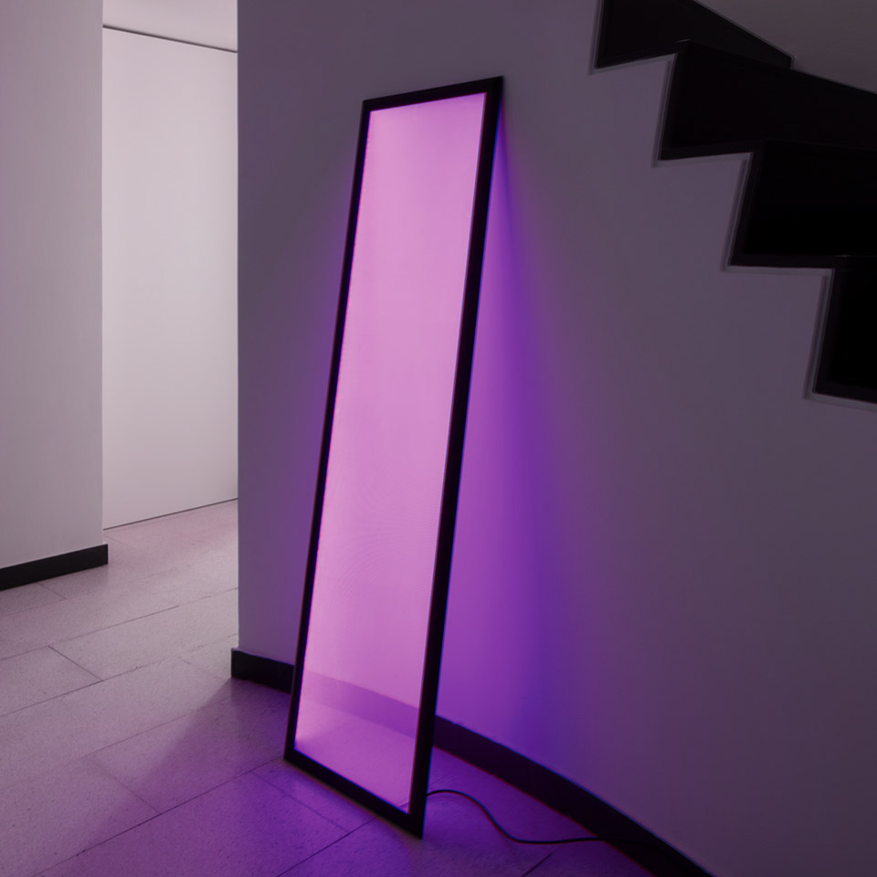 Artemide Discovery Terra floor lamp leaning against wall, emitting vibrant purple light, illuminating surrounding space near staircase