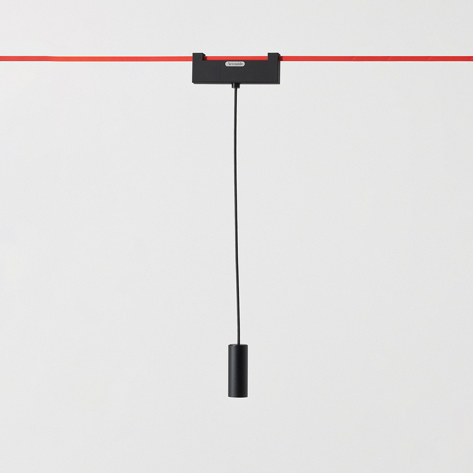 Artemide Funivia pendant lamp: black cylindrical fixture suspended by black cable from a black adapter on a red track rail.