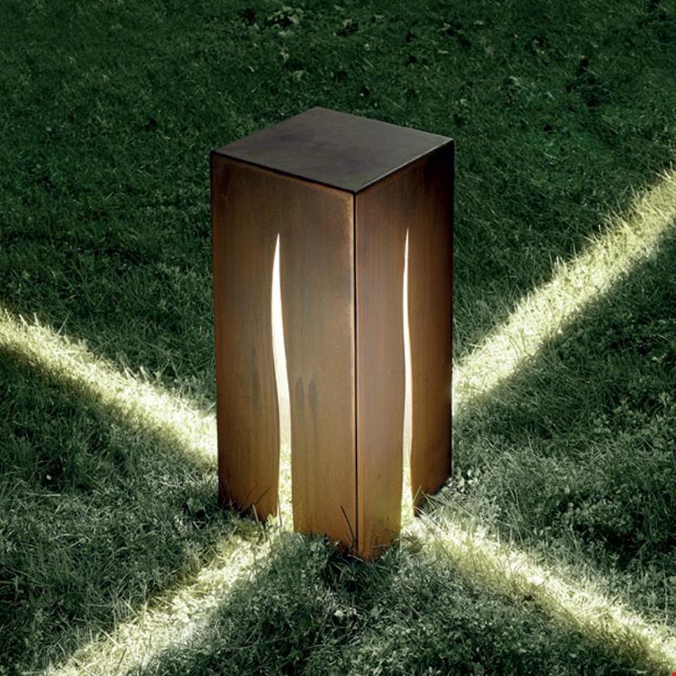 Artemide Granito outdoor bollard light in corten steel with flame-shaped slits casting dramatic light beams on grass