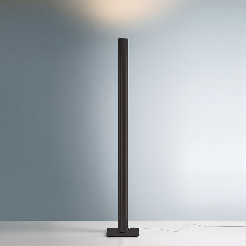 Artemide Ilio floor lamp in black, slim cylindrical column on square base, projecting warm indirect light upward onto the wall