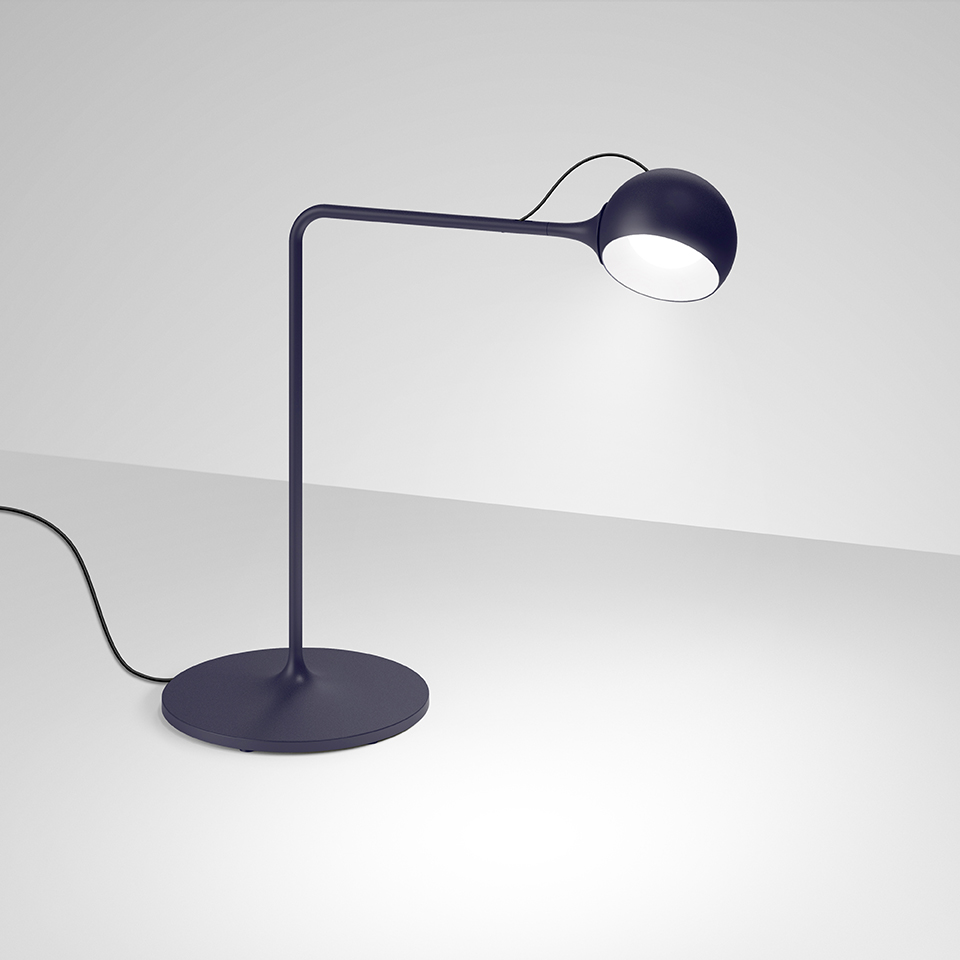 Artemide Ixa desk lamp in dark blue, featuring a round base, curved arm, and adjustable spherical LED head emitting focused light