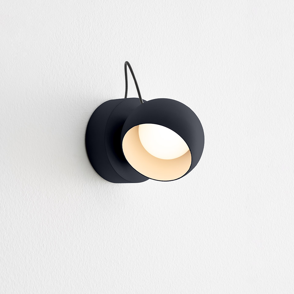 Artemide Ixa wall lamp in black, featuring a round adjustable shade with warm white interior, mounted on a white wall