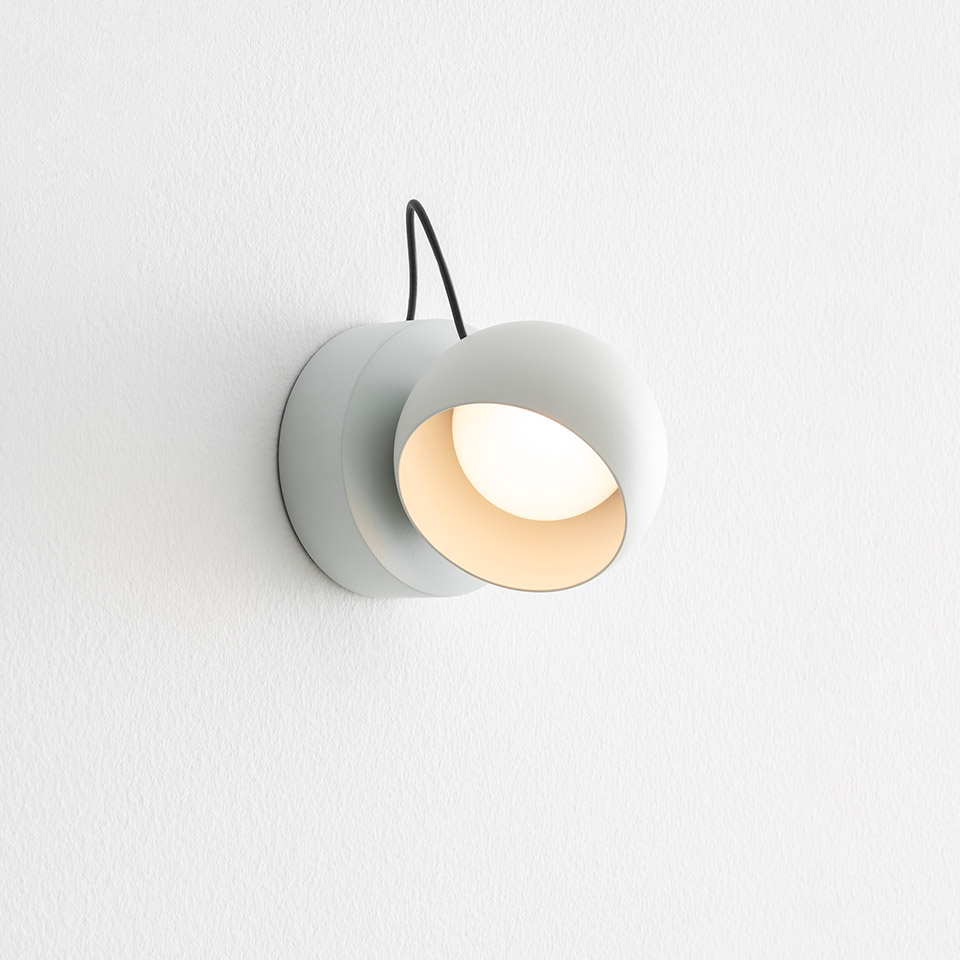 Artemide Ixa wall lamp: compact white cylindrical fixture with rotatable round shade, warm LED light, black cable on white wall