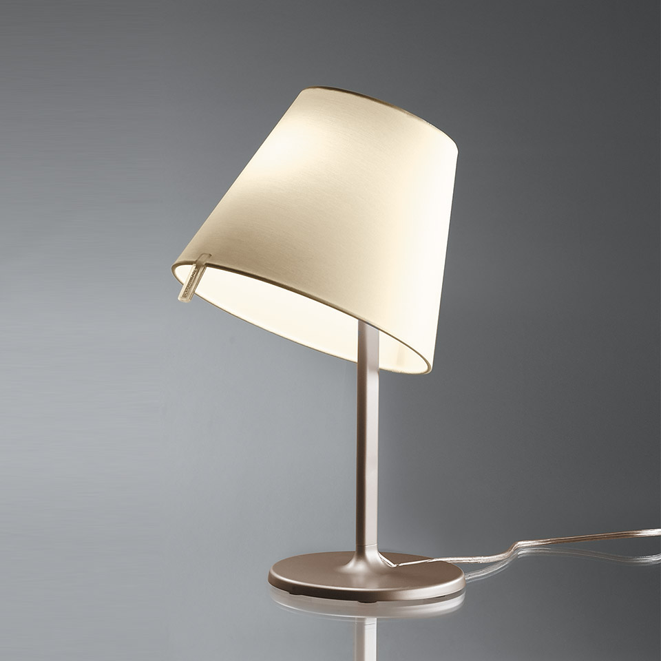 Artemide Melampo Tavolo table lamp with tilted cream fabric shade on a slim brushed aluminum stem and round base, softly lit