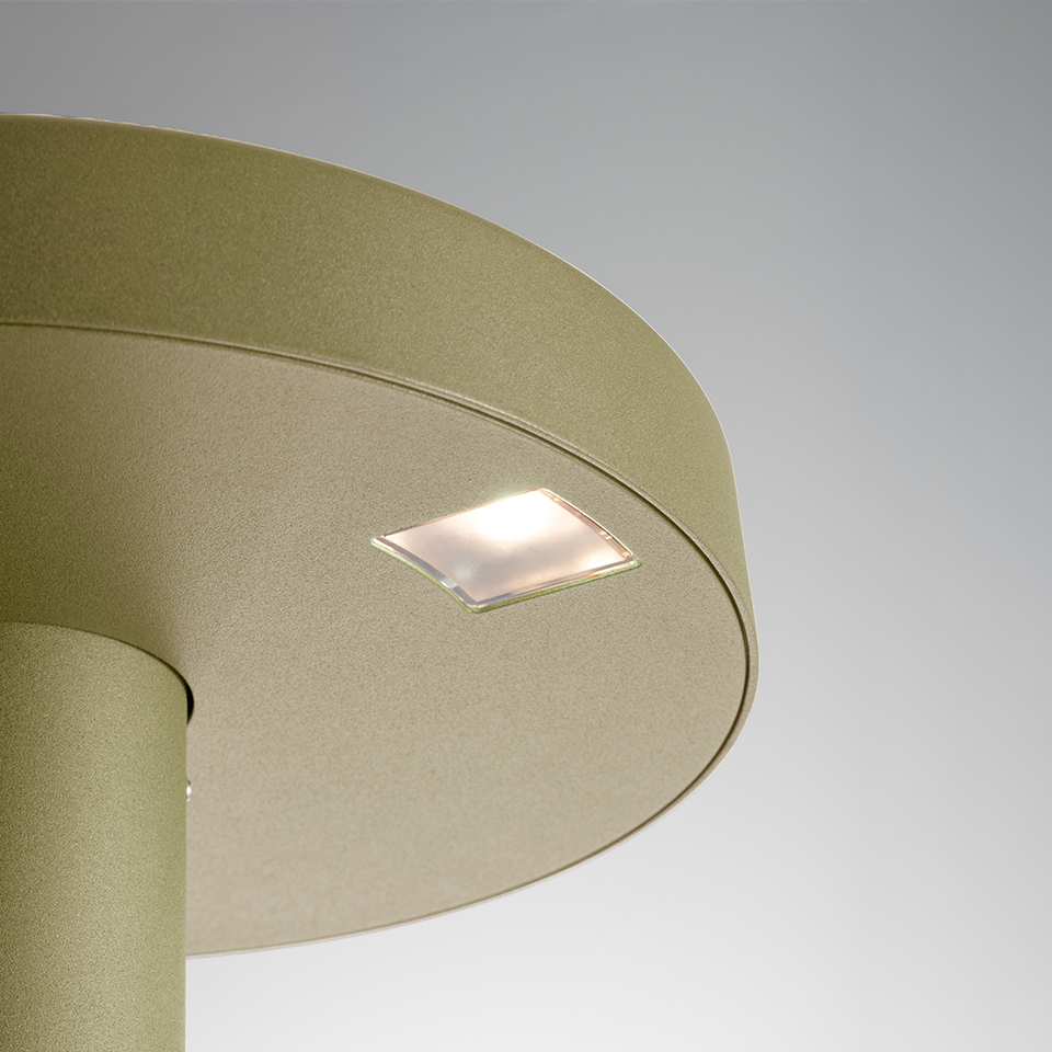 Close-up of Artemide Needoo lamp: gold-green metallic circular shade with small square frosted LED light panel on top surface