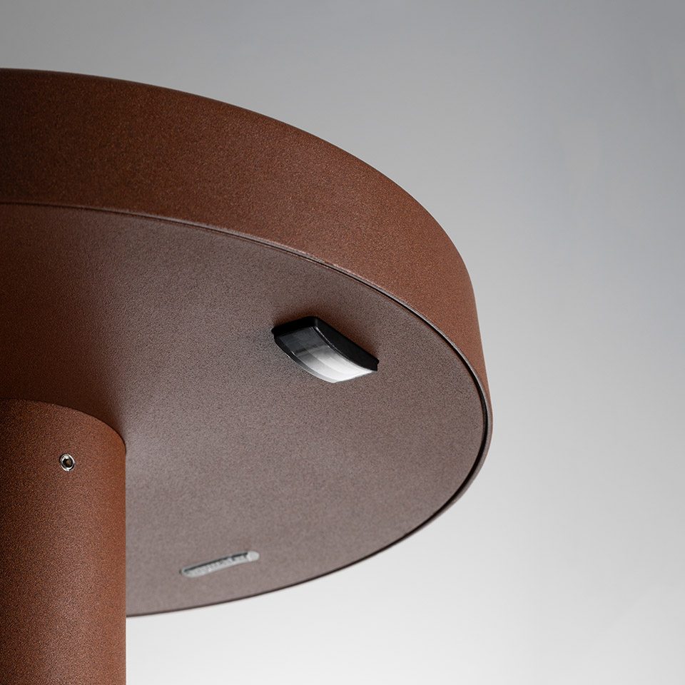 Close-up of Artemide Needoo outdoor lamp base in rust-brown finish, showing motion sensor and charging port on underside