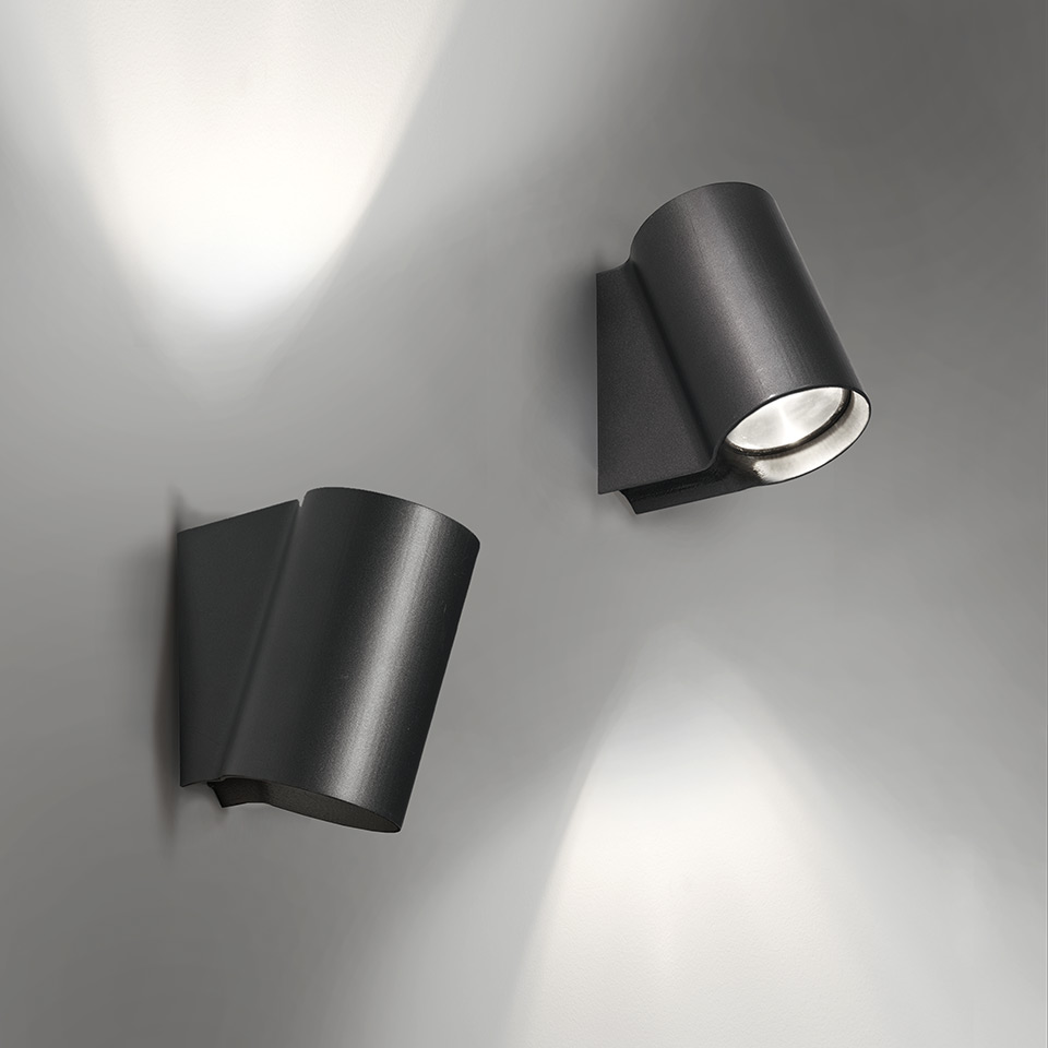 Two Artemide Oblique dark grey wall spotlights: left tilted upward casting wall wash, right angled downward projecting focused beam