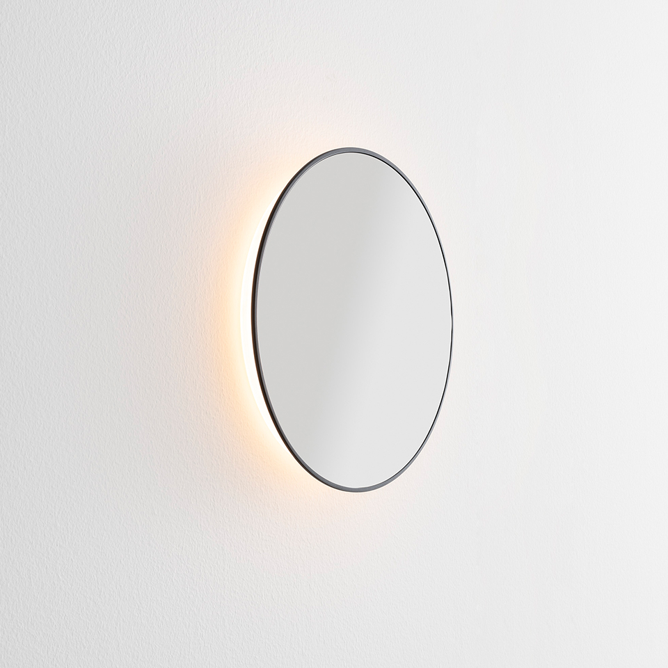 Artemide Se|eS round mirror with thin dark frame and warm LED backlight glowing softly against a white wall