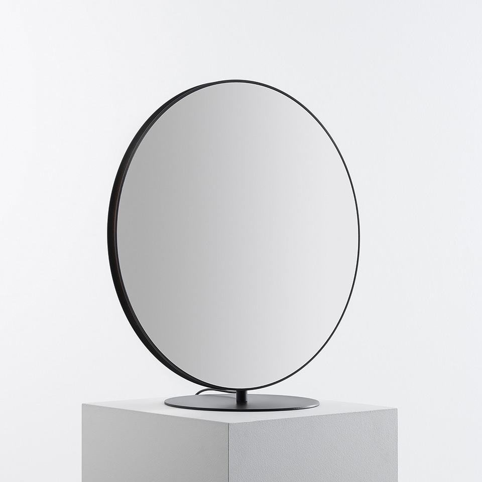 Artemide Se|eS round mirror with thin black metal frame on a circular base, displayed on a white pedestal