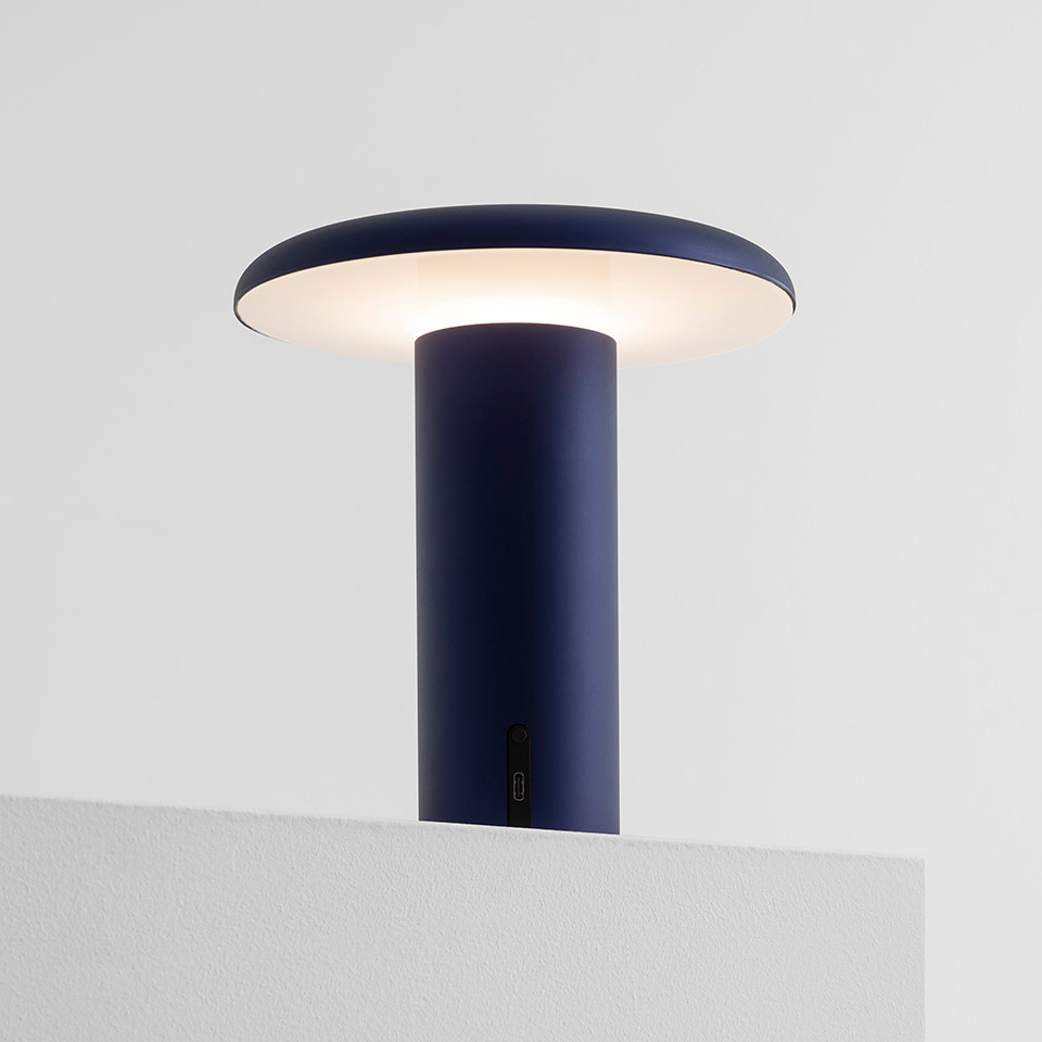 Artemide Takku portable LED table lamp in anodized blue, featuring a cylindrical stem and wide flat diffuser emitting warm light