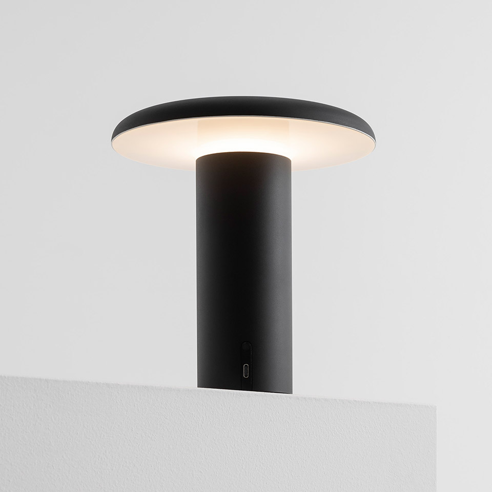 Artemide Takku portable LED table lamp in black, featuring a cylindrical stem and wide flat diffuser emitting warm upward light