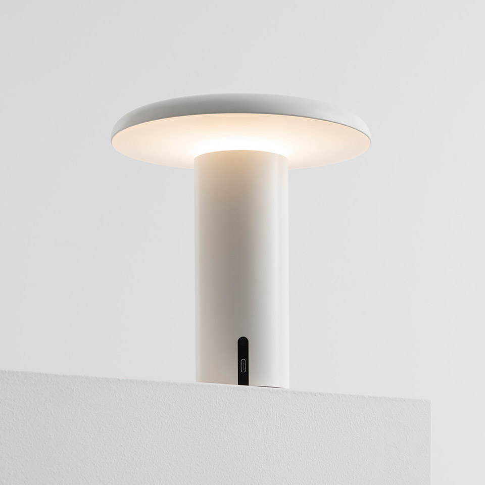 Artemide Takku portable LED table lamp in white, with a flat disc diffuser atop a cylindrical stem, emitting warm ambient light.