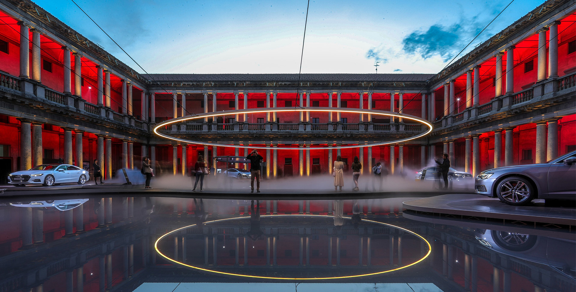 Large luminous ring installation suspended in a historic courtyard, bathed in red light, with cars and visitors reflected on wet floor.