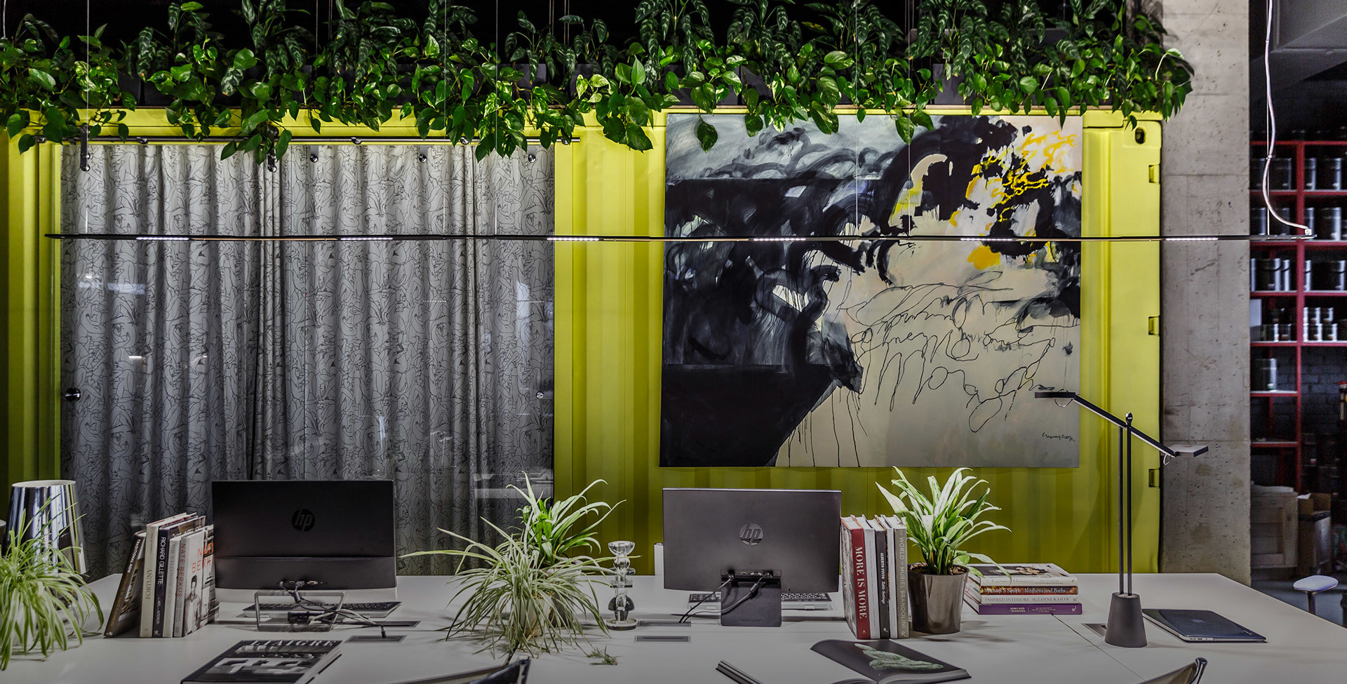 Modern office workspace with Artemide desk lamp, computers, plants, corrugated metal wall, yellow accents, and abstract artwork overhead greenery