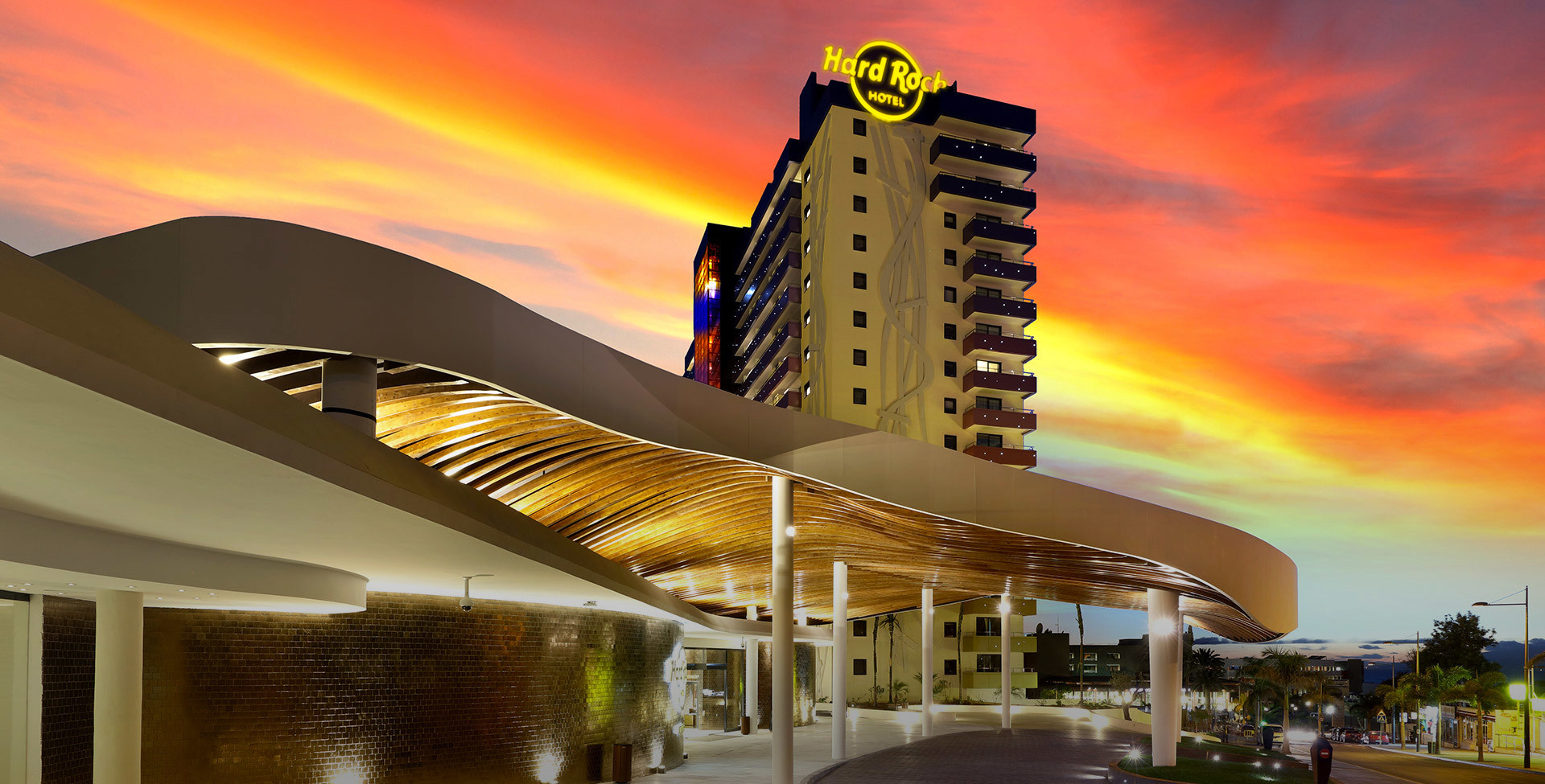 Hard Rock Hotel exterior at dusk, featuring warm golden linear lighting illuminating a curved canopy entrance under a vivid red sunset sky.