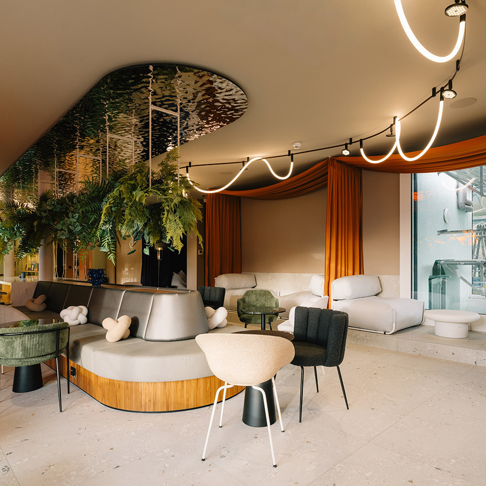 Spacious lounge with upholstered seating, hanging plants, orange curtains, and the Sylt ceiling system with luminous lines.