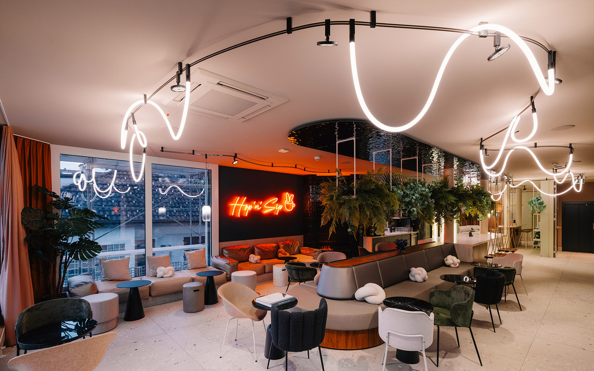 Modern lounge bar with sinuous white LED tube lighting installation on ceiling, neon sign, lush plants, and mixed seating areas