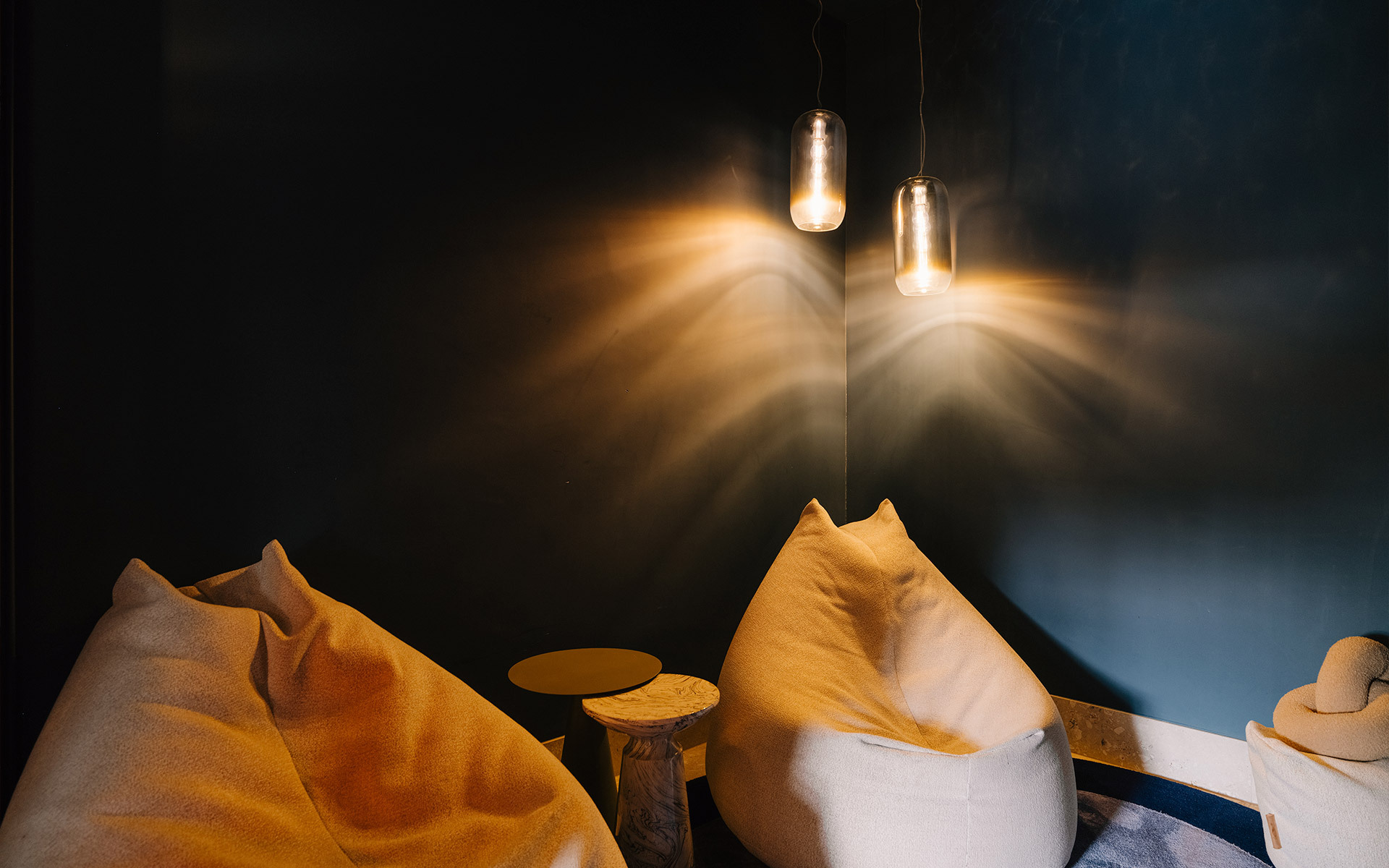 Two cylindrical pendant lamps casting dramatic warm light rays on a dark teal wall above cozy bean bag chairs and a wooden stool.