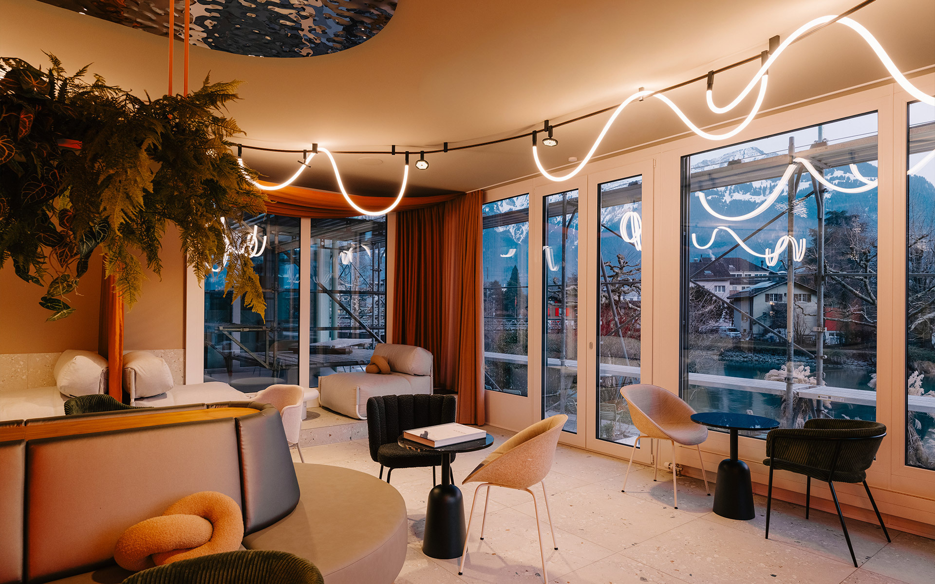 Modern lounge with sinuous white LED neon lighting and warm track lights on ceiling, large windows overlooking a mountain town.