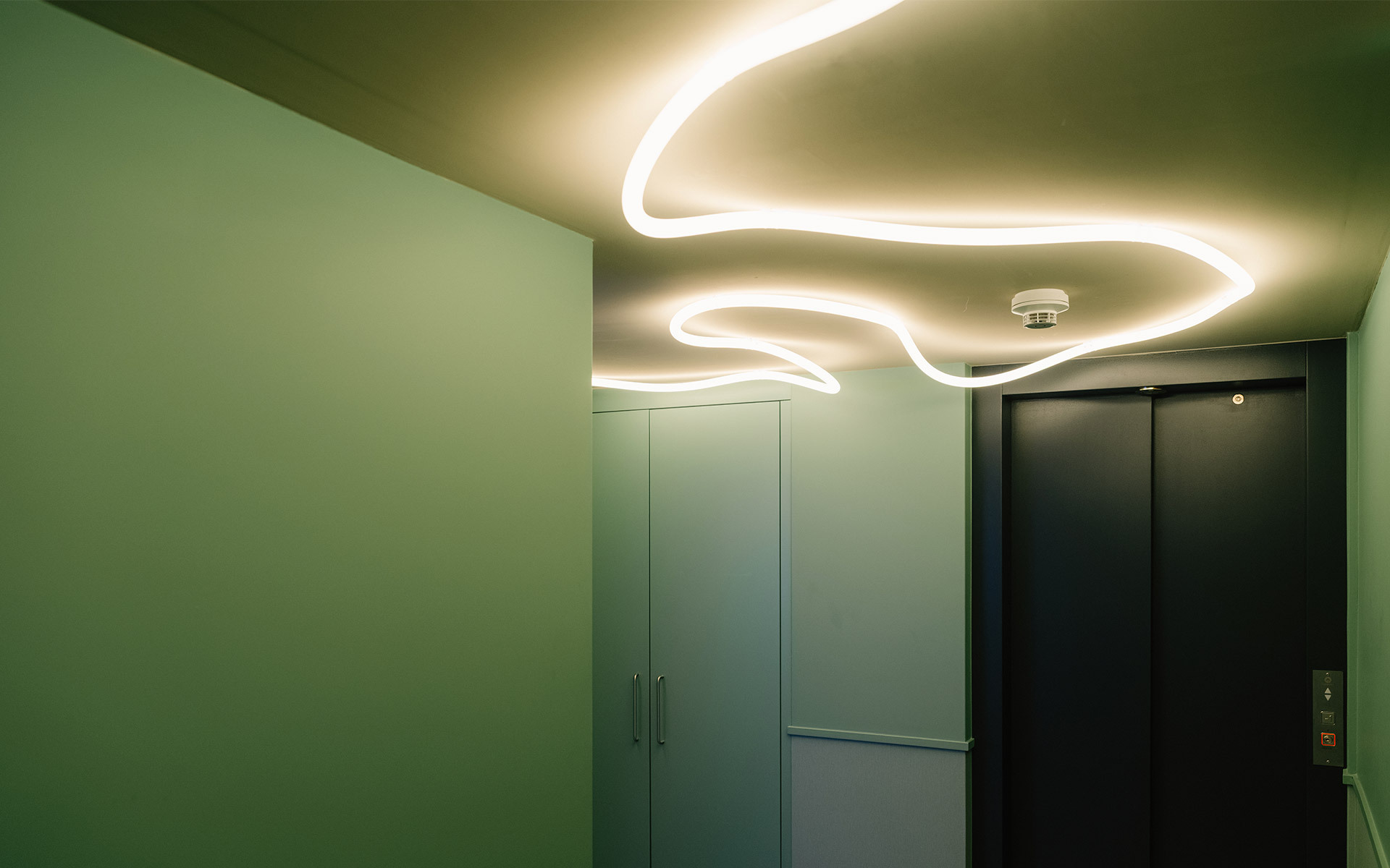 Curved LED light strips forming organic wave shapes on a green ceiling, illuminating a corridor with elevator doors.