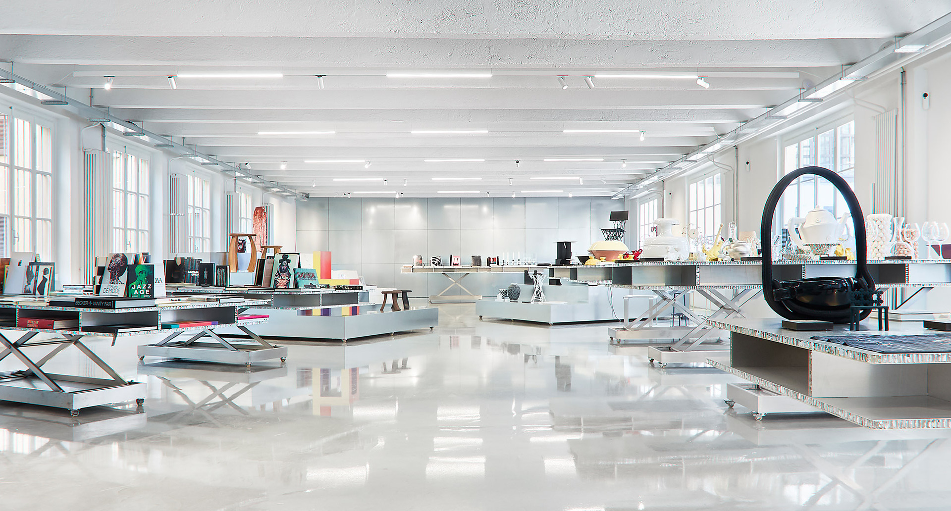 Bright open showroom with white ceilings, recessed lighting, glossy floors, and display tables showcasing various design objects and lamps.