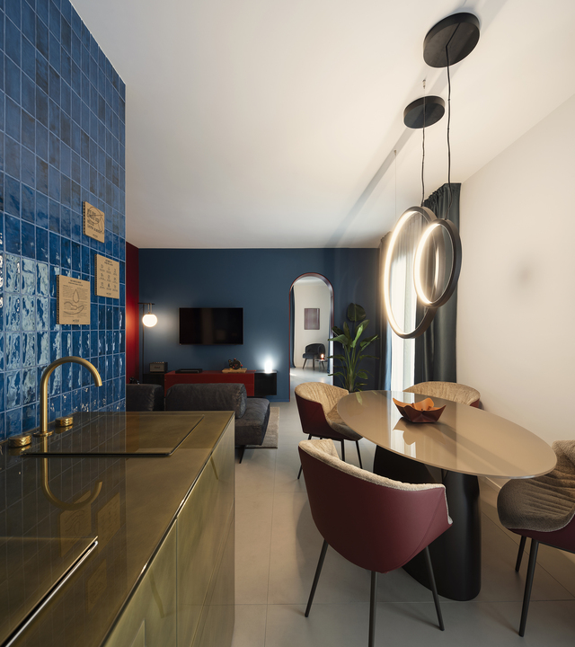View of the living room with the dining table illuminated by “O” suspension lights; in the background, the sofa and TV can be seen, while the kitchen appears in the foreground.