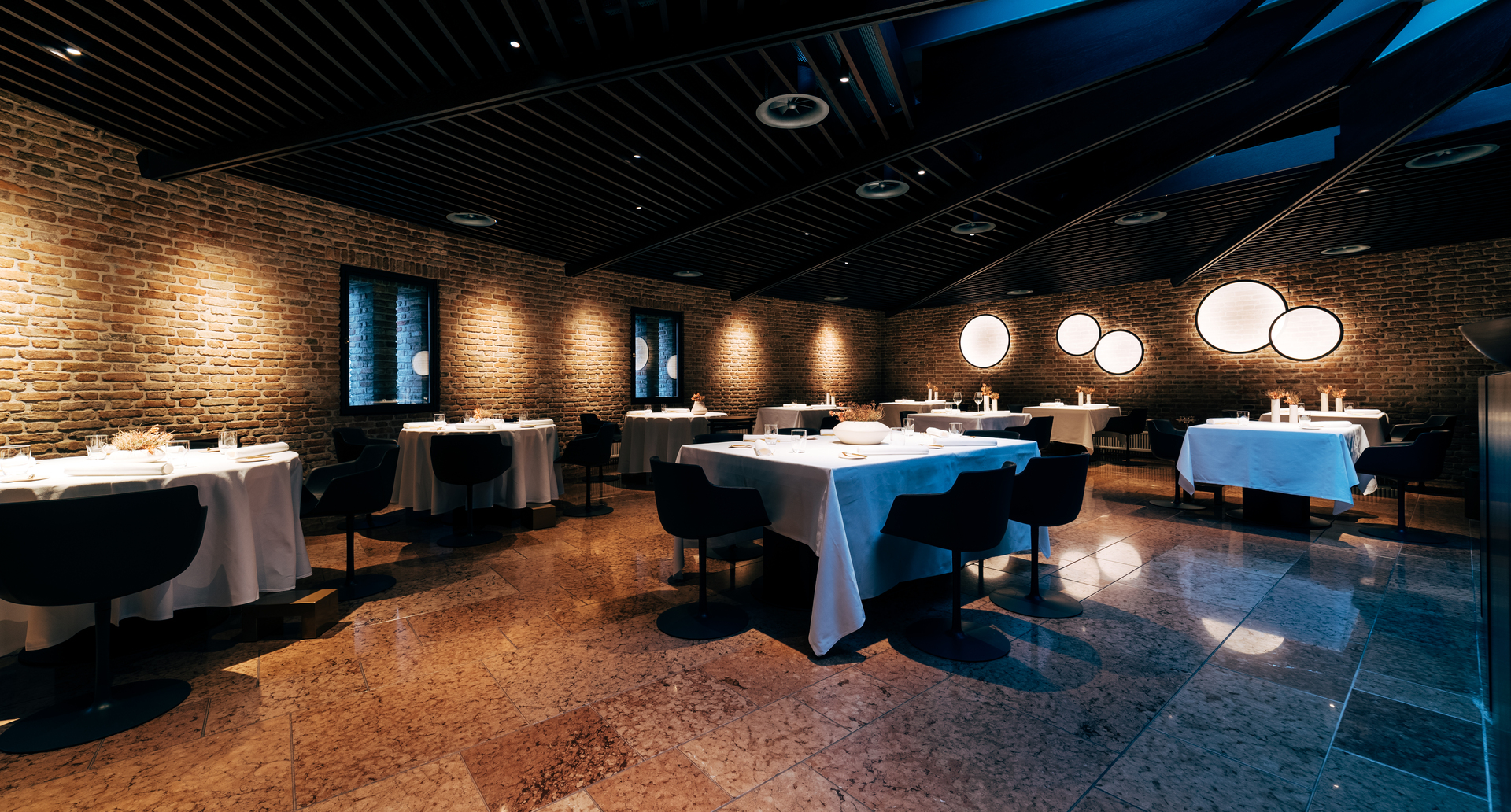 Elegant restaurant dining room with exposed brick walls, marble floor, white-clothed tables, and circular Artemide wall lights creating warm ambiance.