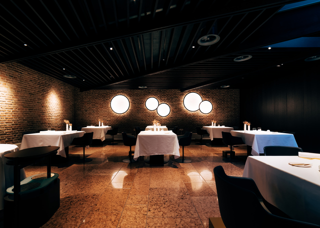 Elegant restaurant dining room with dark ceiling, brick walls, white-clothed tables, candles, and circular wall-mounted luminaires providing warm ambient light.