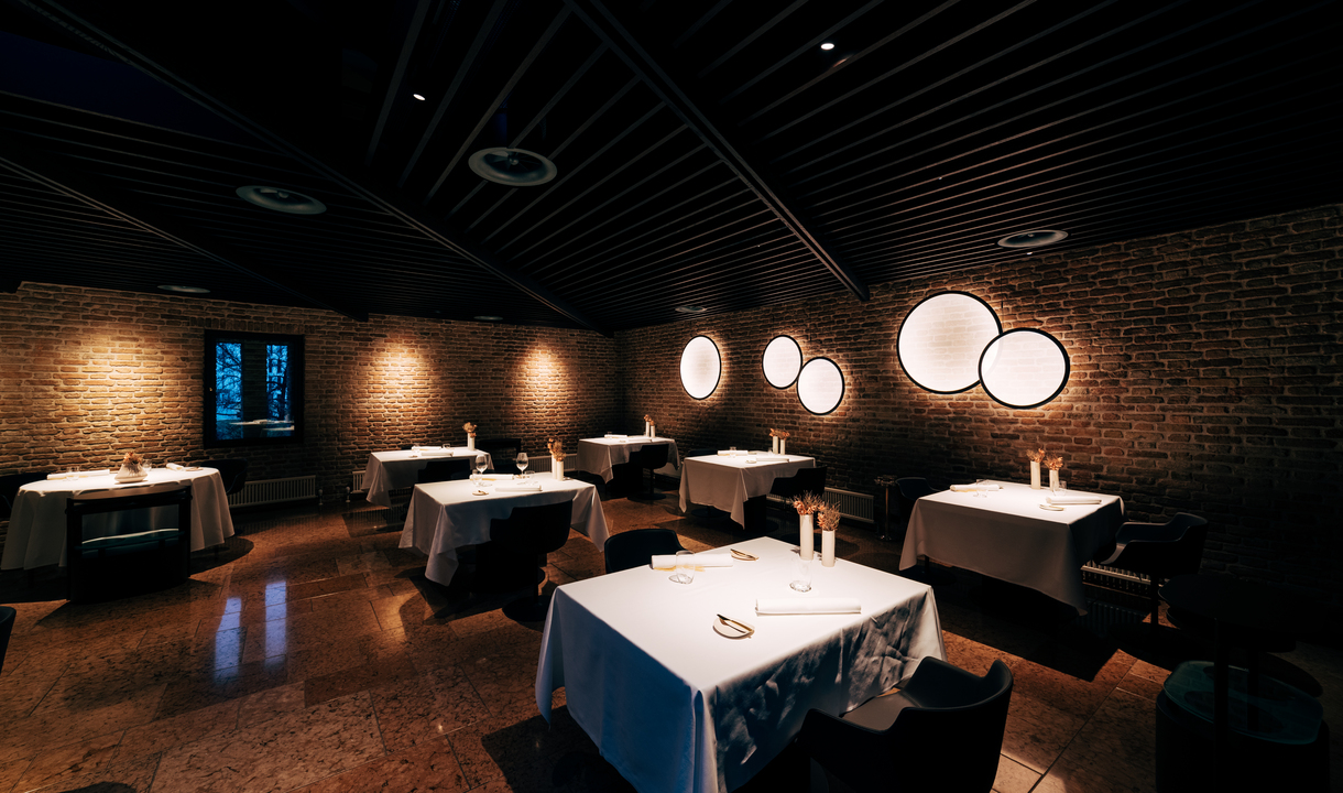 Elegant restaurant dining room with brick walls, dark ceiling, white-clothed tables, and circular Artemide wall lights creating warm ambient lighting.