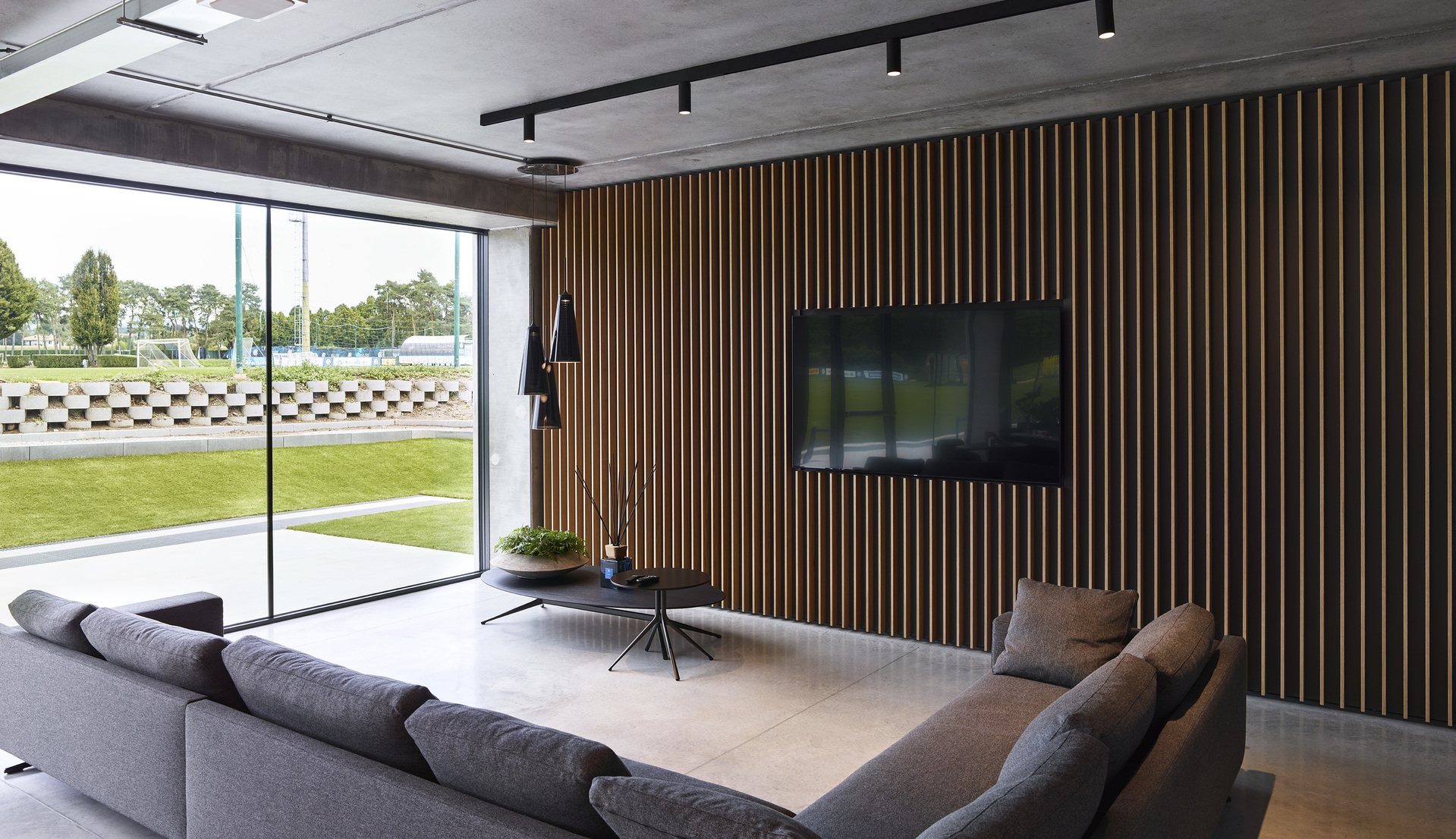 Modern living room with gray sectional sofa, wood-slat accent wall, wall-mounted TV, and Artemide track lights on concrete ceiling