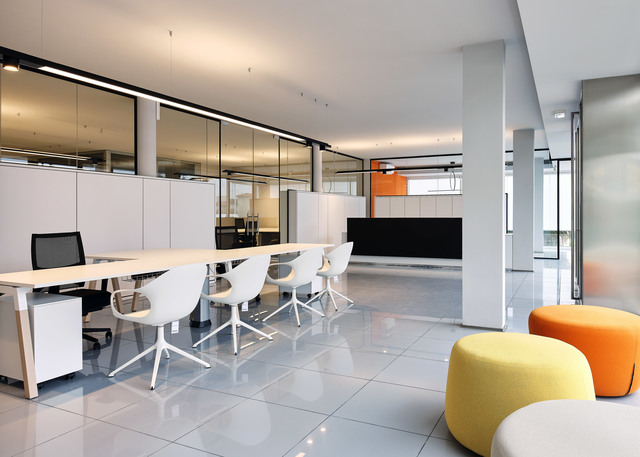 Modern open office with Artemide linear ceiling lights, white workstations, glass partitions, and colorful lounge seating in yellow and orange