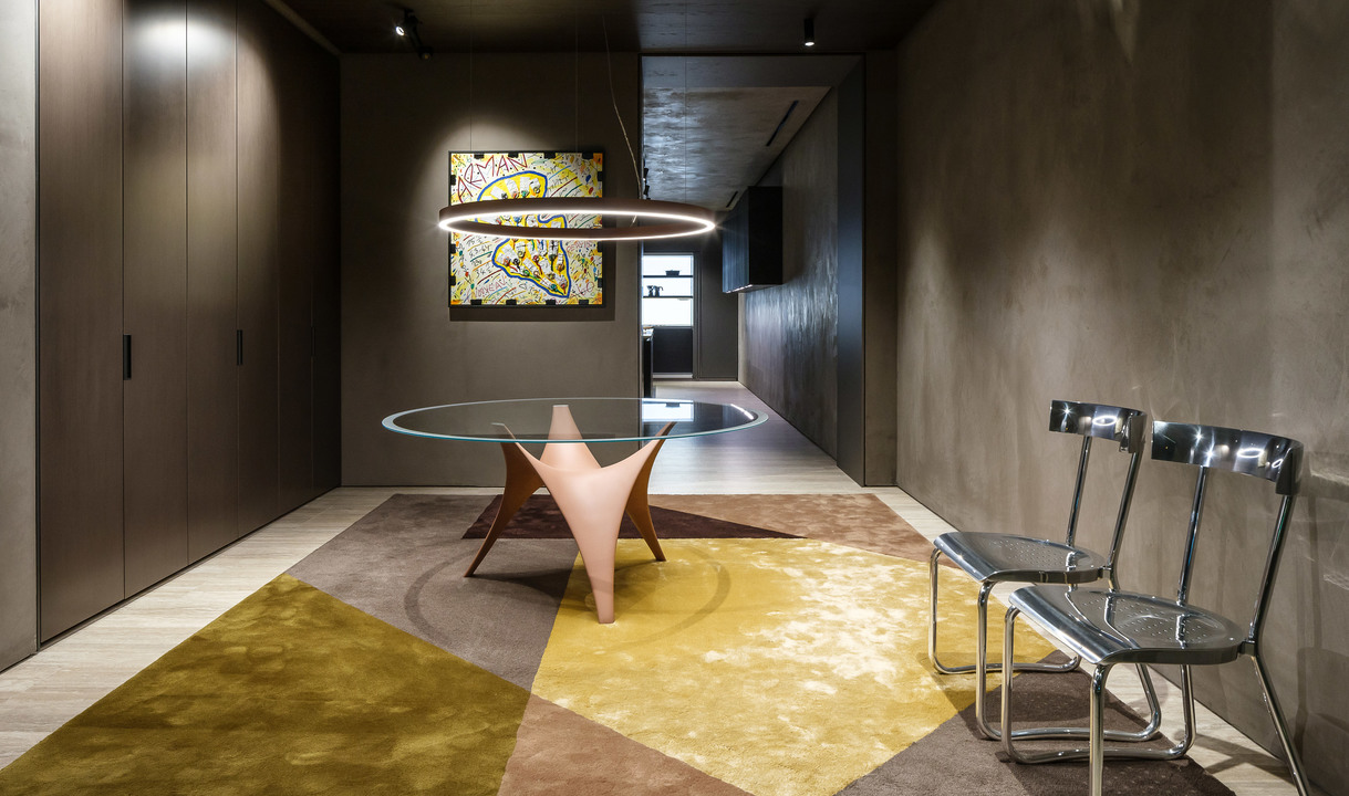 Artemide circular ring pendant lamp over a glass dining table on a geometric rug, in a modern dark interior with artwork.