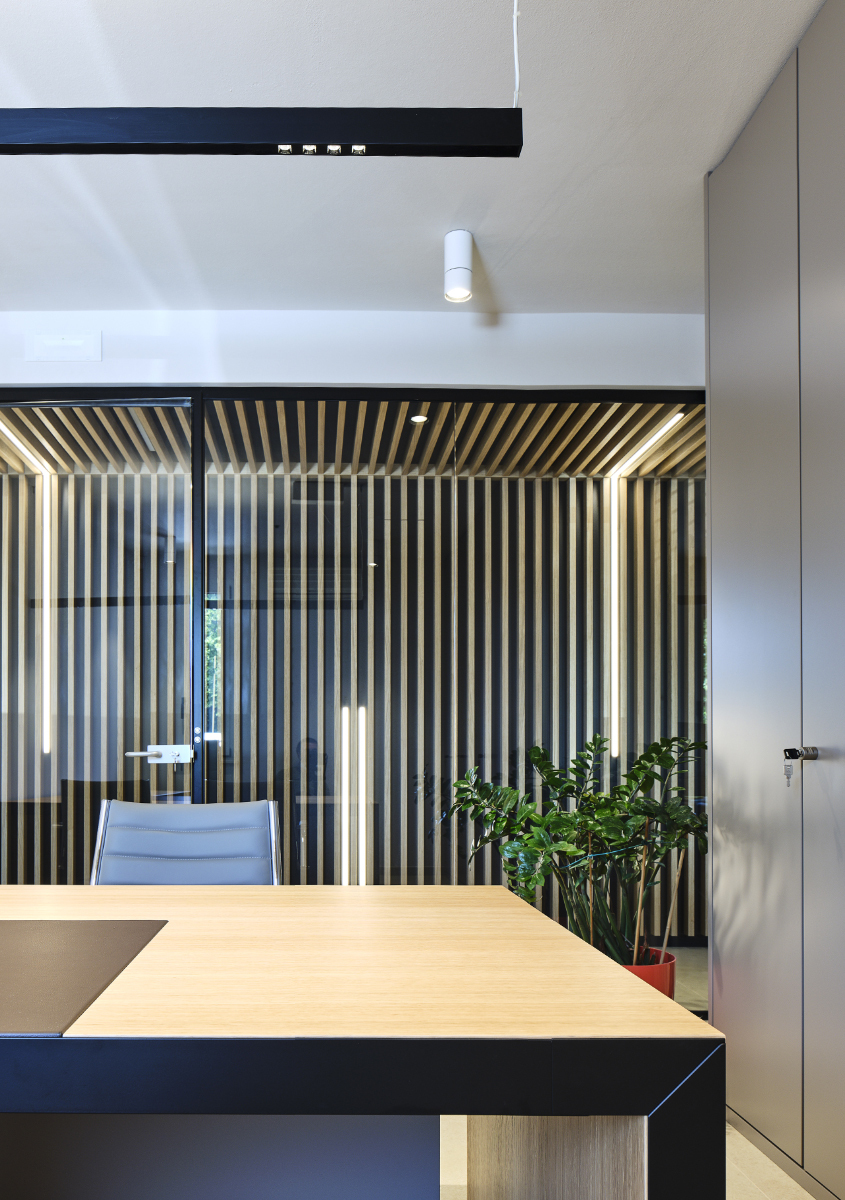 Modern office with wooden desk, black linear pendant lamp, white cylindrical spotlight, and illuminated slatted wood partition wall.