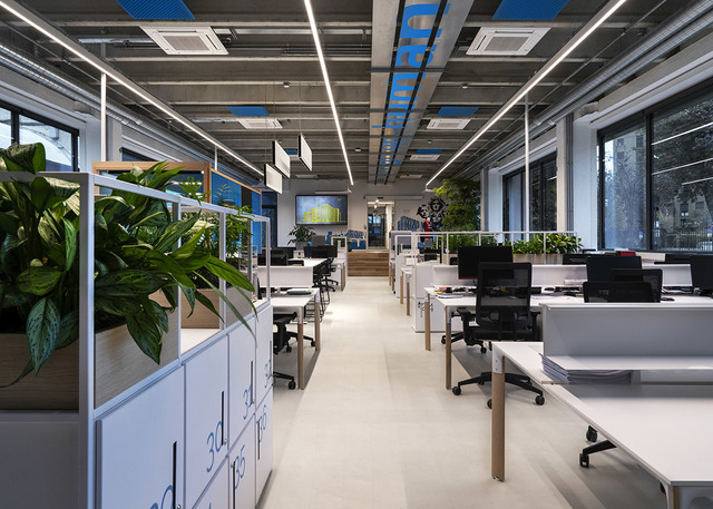 Modern open-plan office with Artemide linear LED ceiling lights illuminating workstations, white desks, ergonomic chairs, and green plants.