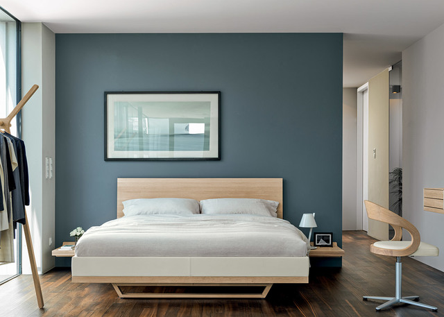 Modern bedroom with teal accent wall, light wood bed frame, white bedding, small bedside lamps, and a sculptural chair