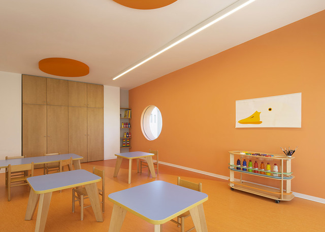 Bright children's classroom with orange walls, small tables and stools, lit by a recessed linear LED ceiling light.