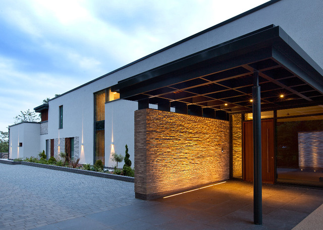 Modern home entrance at dusk with warm uplighting on stone wall, recessed ceiling lights under canopy, and white wall-mounted fixtures
