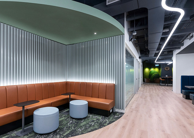 Modern office lounge with curved orange leather seating, lit backwall panels, and zigzag LED linear ceiling lights in the corridor.