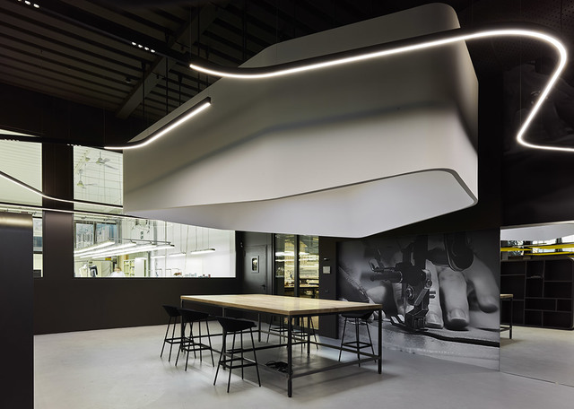 Modern workspace with a sculptural white ceiling installation featuring integrated LED linear lighting above a long wooden table with black stools.