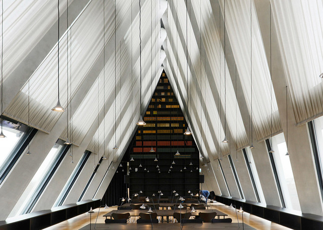 Grand library interior with soaring triangular ceiling, pendant lights hanging over long reading tables, colorful bookshelves in background