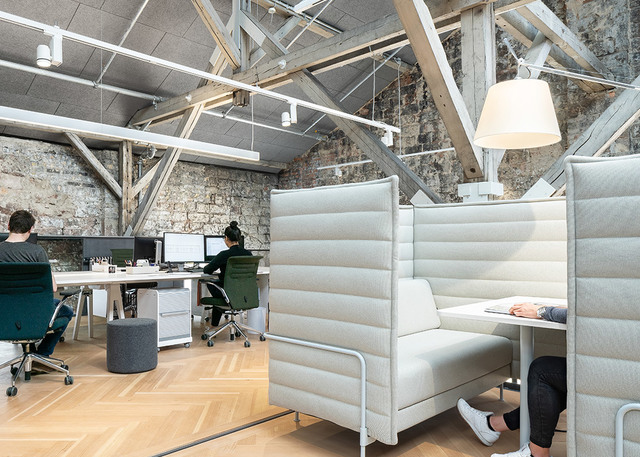 Modern office with exposed stone walls, wooden trusses, workstations, and a white pendant lamp over a high-back upholstered booth seating area.