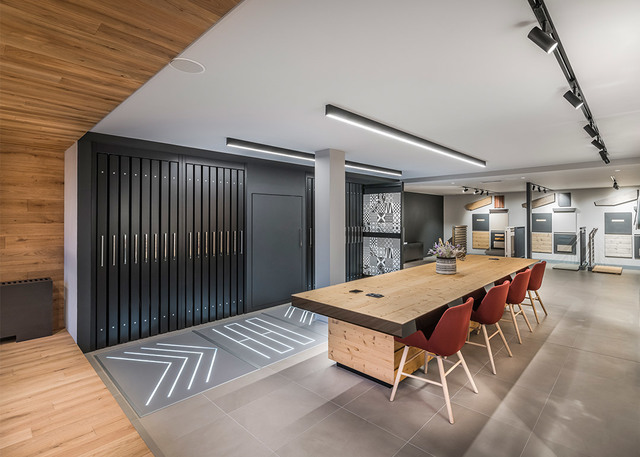 Modern showroom with linear ceiling lights, track spotlights, a wooden meeting table with red chairs, and decorative floor lighting.
