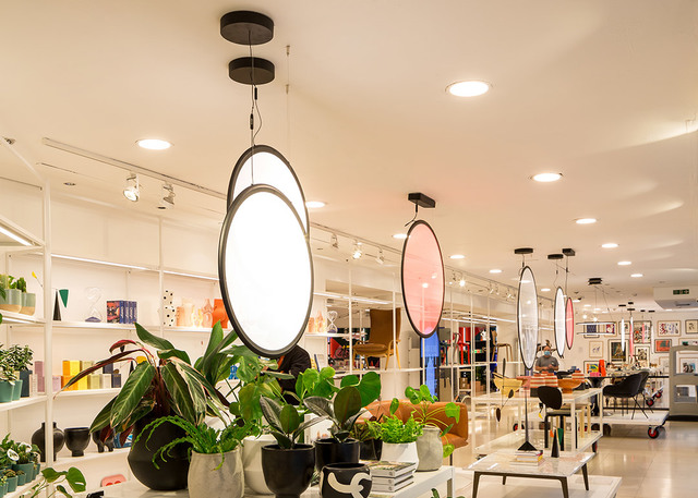Retail interior with Artemide oval pendant lamps in white and pink tones, suspended from black frames, above shelves with plants and decor.