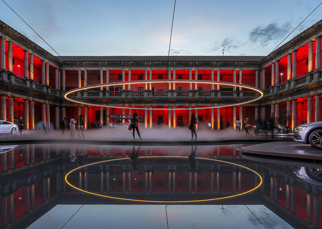 Large glowing circular ring light suspended over a misty courtyard, reflected in a pool, with a red-lit neoclassical building at dusk