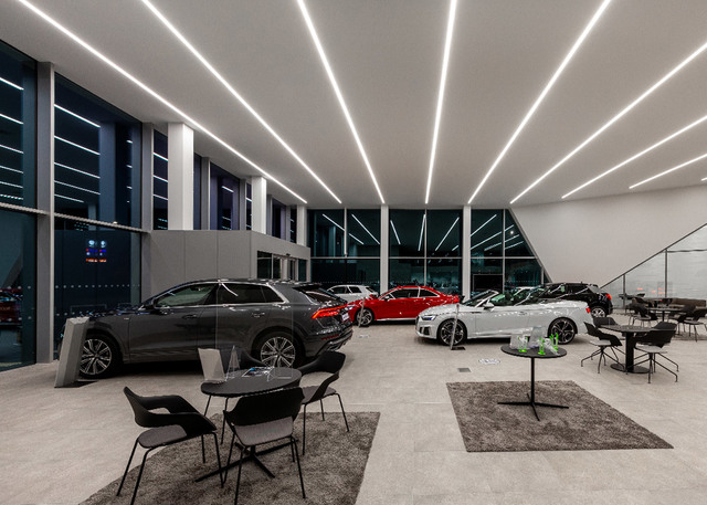 Modern car showroom with linear LED ceiling lights illuminating luxury vehicles on a polished concrete floor.