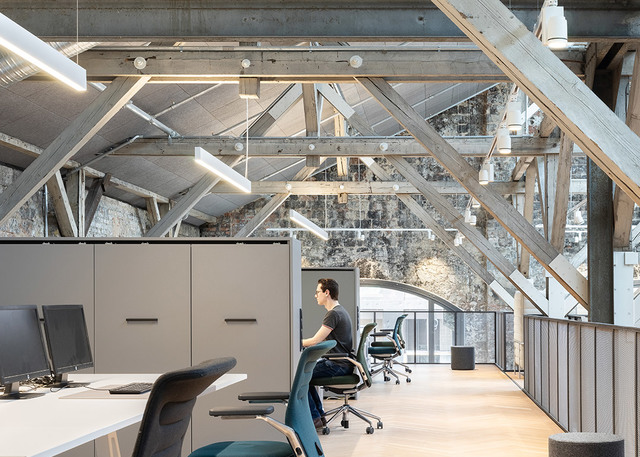 Modern office workspace with Artemide linear pendant lights under exposed wooden roof trusses, workers at desks with teal ergonomic chairs