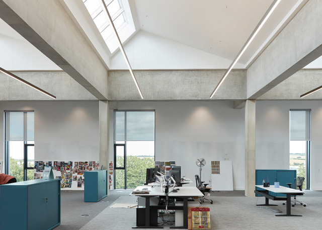 Modern open-plan office with high concrete ceilings, skylights, linear LED ceiling lights, workstations with monitors, and large windows overlooking greenery.