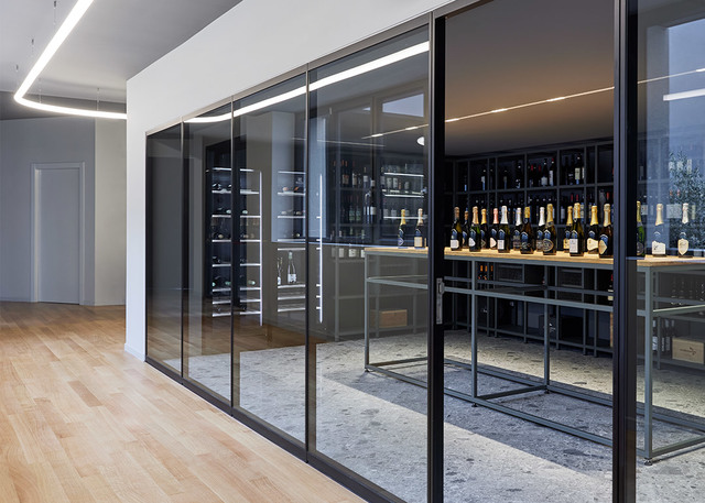 Glass-enclosed wine cellar with dark shelving displaying bottles, illuminated by a recessed linear LED ceiling light strip.