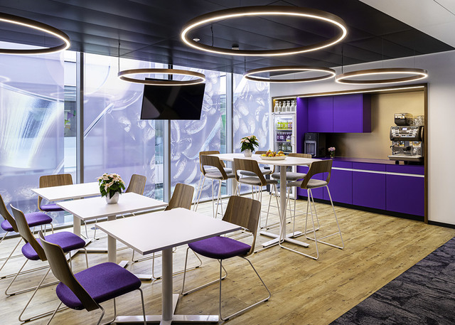 Modern office breakroom with Artemide circular ring pendant lights illuminating white tables, purple chairs, and a purple kitchenette.