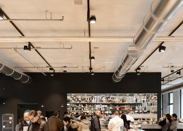 Busy café interior with exposed industrial ceiling, silver HVAC ducts, and black track-mounted Artemide spotlights illuminating a crowded bar area.